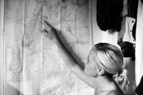 A young woman in profile points at a large, crumpled map on a wall, her blonde hair tied back.