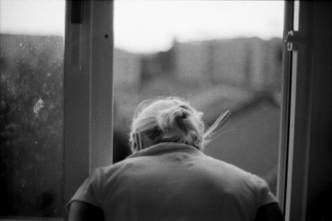 A black and white photo of an older person seen from behind, looking out a window at a blurry exterior.