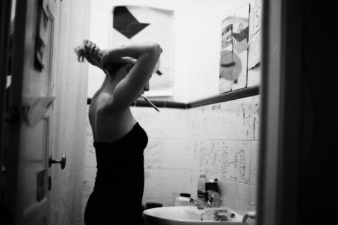 Black and white photo of a woman in a bathroom, seen from the side, brushing her teeth and tying her hair.