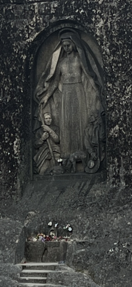 Grayscale image of a stone religious monument featuring a large robed figure, smaller figures, and a flower-adorned altar…