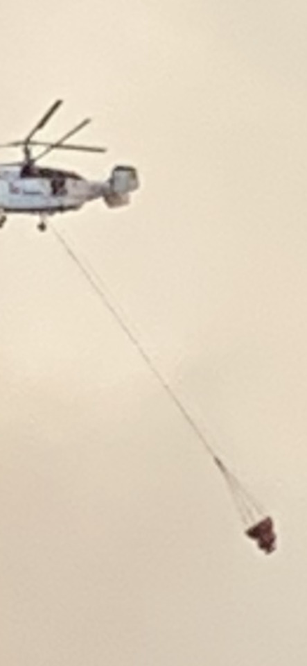 A helicopter flies through a hazy sky, carrying a large water bucket suspended by a long cable.