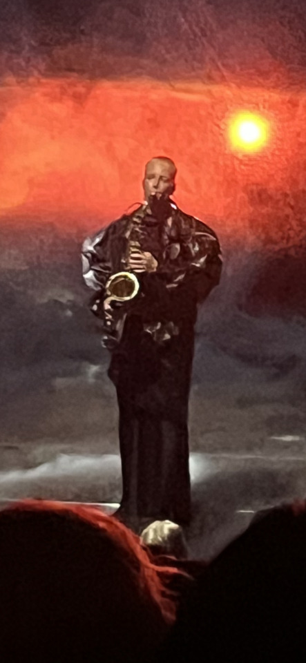 A bald man in a dark robe plays a saxophone on stage against a dramatic orange and dark sky with a glowing sun.