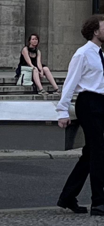 A woman sits on stone steps in the background while a man in a white shirt and tie walks past in the foreground.