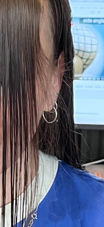 A close-up of a person's side profile with wet, dark hair, silver hoop earrings, and a blue shirt, with a bright screen in…