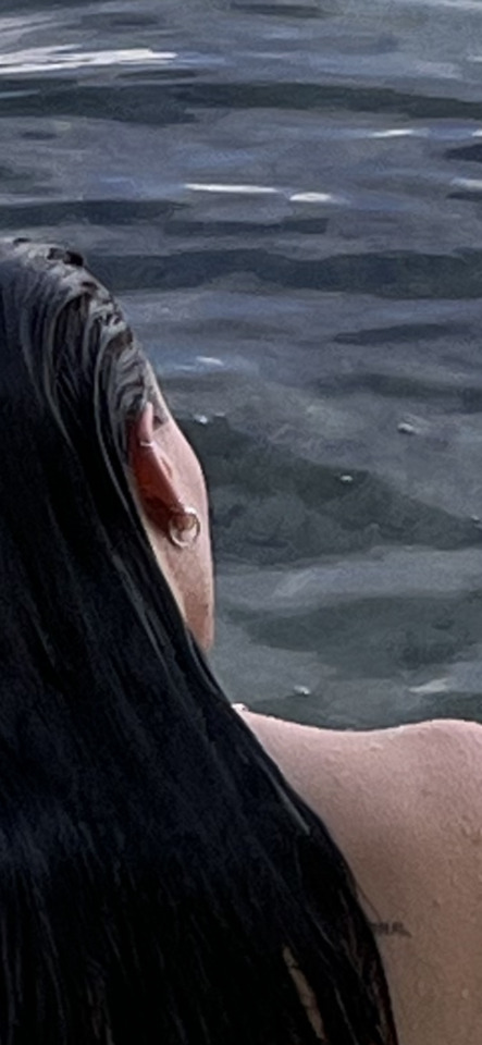 Close-up of a person with wet dark hair and an earring, looking out over calm, dark water.