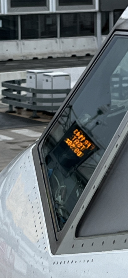 Close-up of an airplane cockpit window reflecting an orange digital display, with the aircraft fuselage and airport…