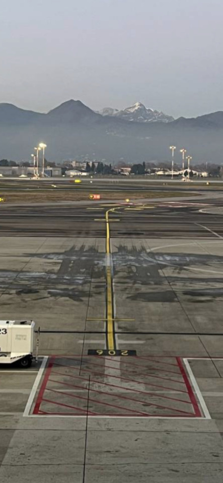 An airport tarmac and runway stretch towards distant hazy mountains, some snow-capped, under a soft, diffused light.