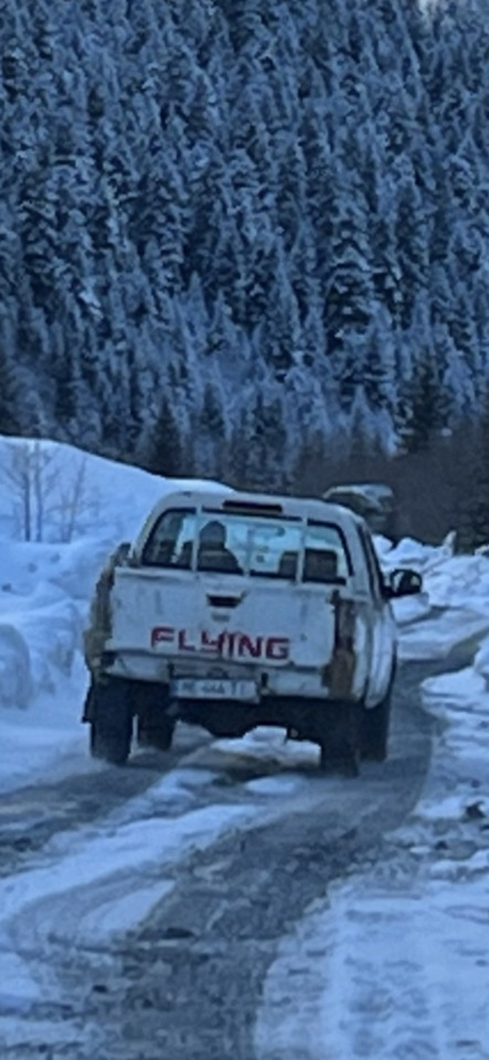 White pickup truck with "FLYING" on its tailgate drives on a snowy road through a winter forest.