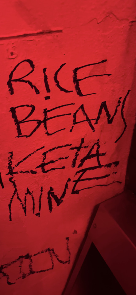 Black graffiti on a red-lit wall reads "RICE," "BEANS," "KETAMINE," and "WINE."