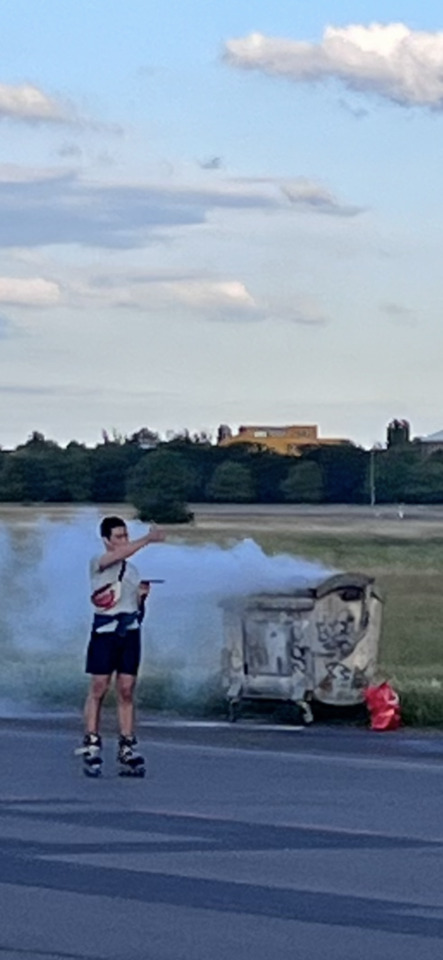 A person on rollerblades points at a graffiti-covered dumpster emitting a large cloud of smoke on the side of a road.