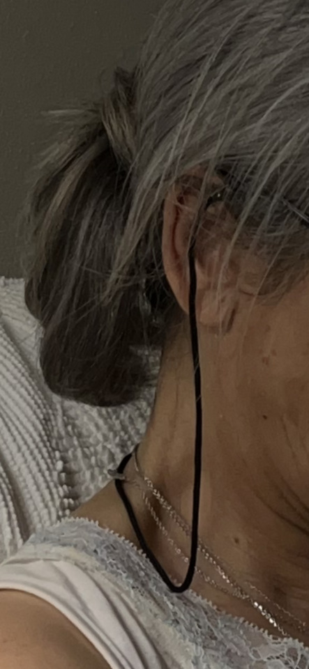 Close-up of a person's side profile, showing grey hair, an ear, a black cord, and multiple silver necklaces.