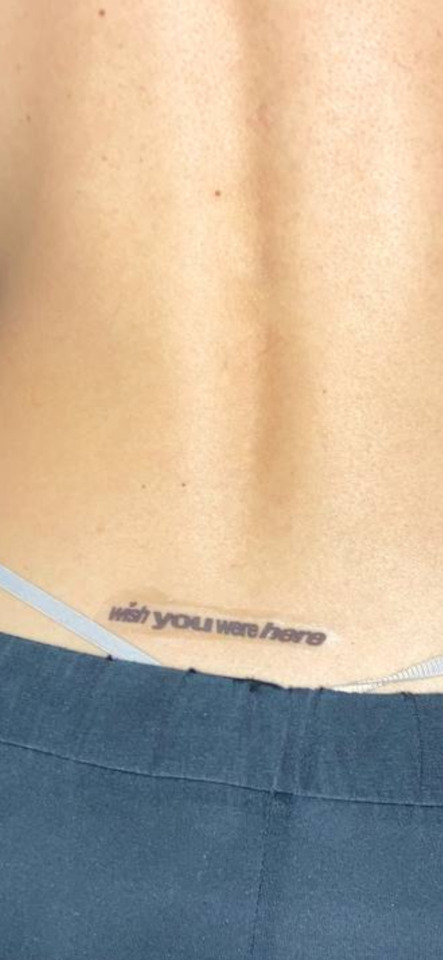 Close-up of a person's lower back with a "wish you were here" tattoo above dark blue pants.