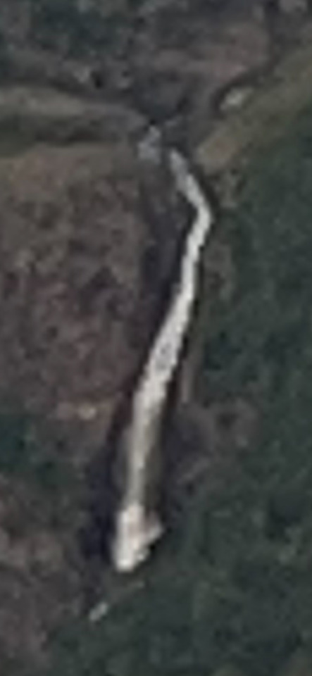 Aerial view of a long, pale, winding feature resembling a river or path, set against a dark, textured landscape.