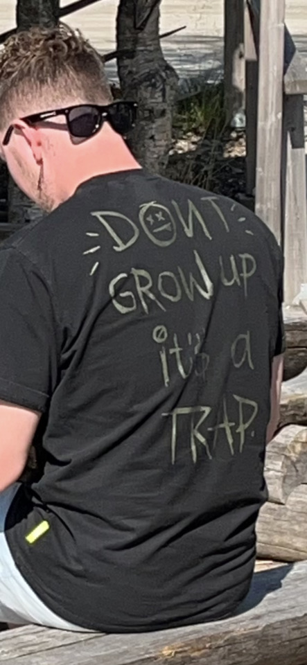 A man from behind wearing a black t-shirt that says "DON'T GROW UP IT'S A TRAP" with a smiley face.