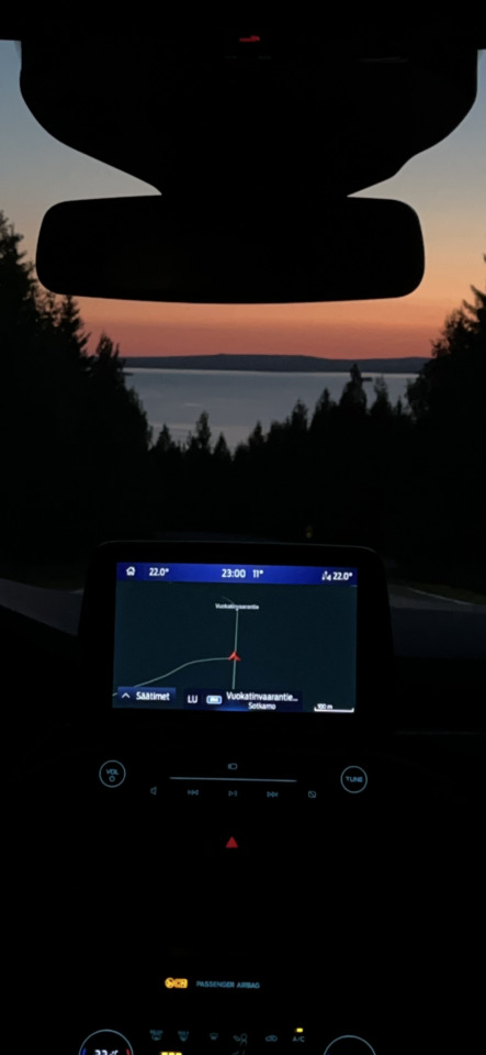 A view from inside a car at dusk, showing a navigation screen and a sunset over a lake framed by trees.