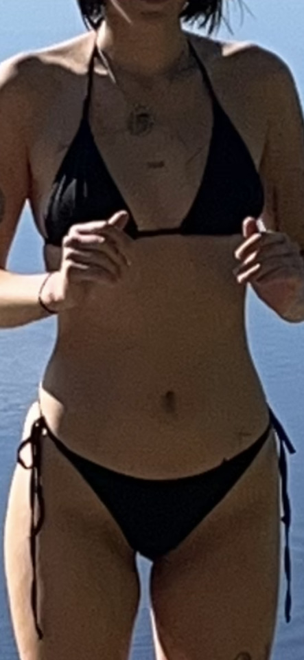 A person in a black bikini stands against a blue water background, hands near their chest, under bright sunlight.
