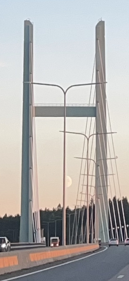 A cable-stayed bridge at twilight with two tall towers, cars on the road, and a full moon visible between the towers.