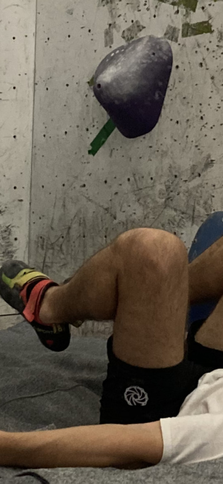 A person's legs and lower torso, wearing climbing shoes and shorts, resting on a mat in a climbing gym.