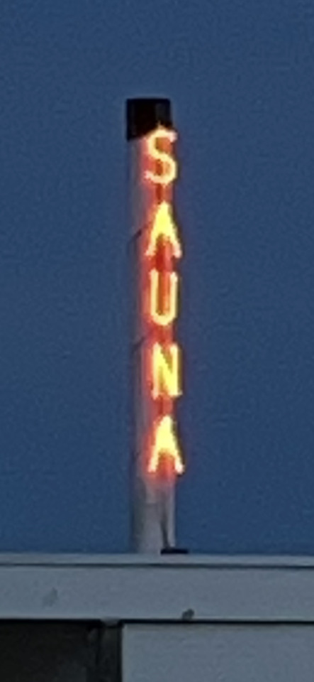 A vertical neon sign glowing orange against a dark blue sky, spelling out "SAUNA".