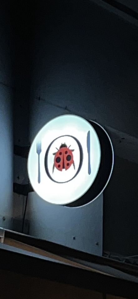 A circular, illuminated sign shows a red ladybug on a white plate with a blue fork and knife against a dark background.