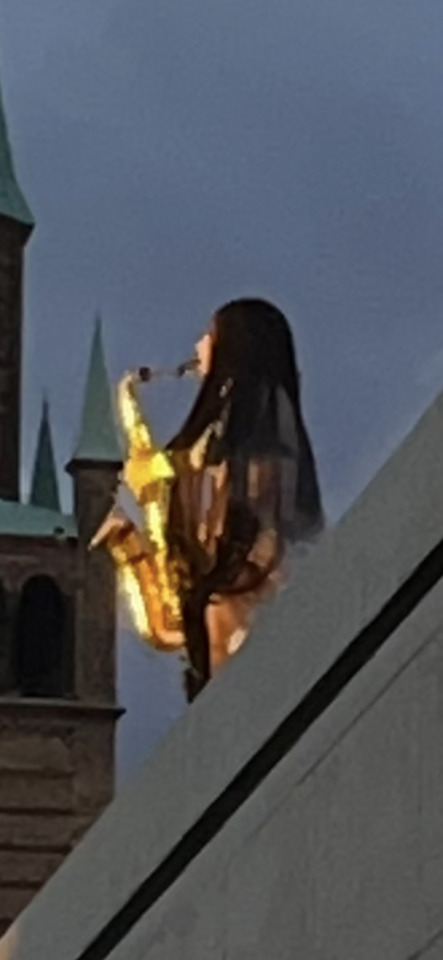 A person with long dark hair plays a golden saxophone outdoors, with old building spires in the background.