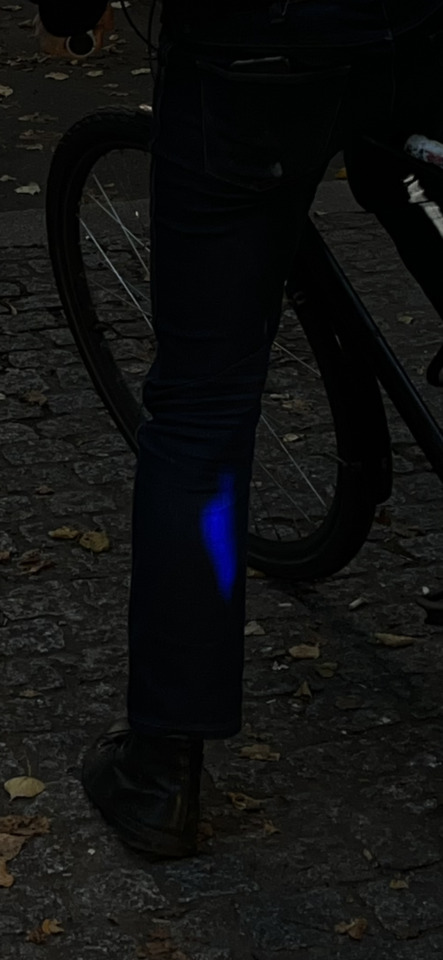 A person in dark clothing stands next to a bicycle in a dim setting, with a bright blue light on their lower leg.