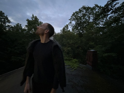 Man in glasses and dark clothes looks up at a cloudy sky from a dark rooftop surrounded by trees.
