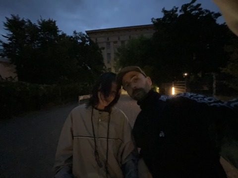Two people pose for a low-light photo outdoors at night, with a large building and trees in the dark background.