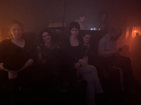 A group of people sit in a very dimly lit room with a warm, reddish-orange glow, some looking at the camera.