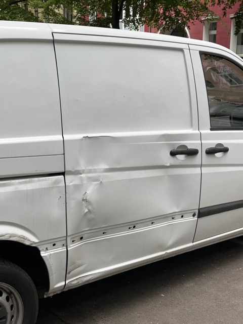 White van with severe side body damage, including large dents and missing trim, parked outdoors in daylight.