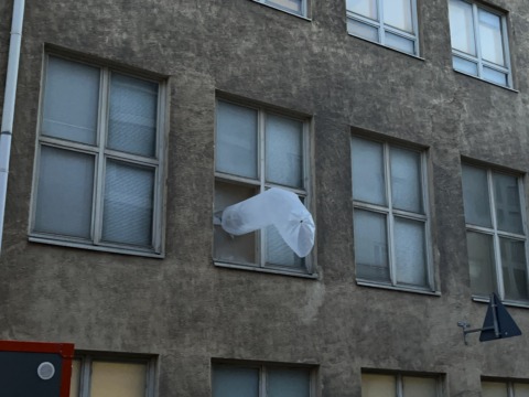 A weathered grey building with many windows, one of which has a large white translucent object billowing out.