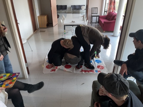 People play Twister on a white tiled floor in a brightly lit room, while others watch.