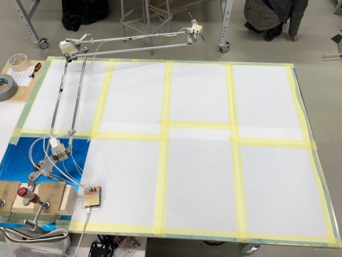A mechanical arm setup is clamped to a large white board taped into a grid, with electronics and tools nearby.