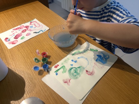 A child in a striped shirt paints at a wooden table with small pots of paint, a bowl of water, and two painted papers.