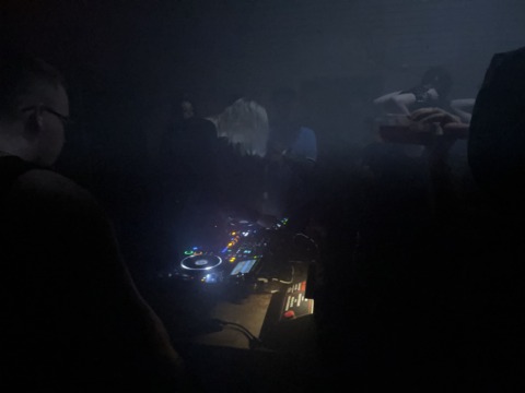 A DJ plays music in a dark, smoky club with glowing equipment and silhouetted people.